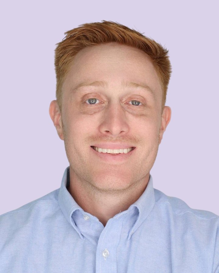 Jonah Hancock smiling, wearing a light blue button-down shirt, in front of a lavender backdrop.